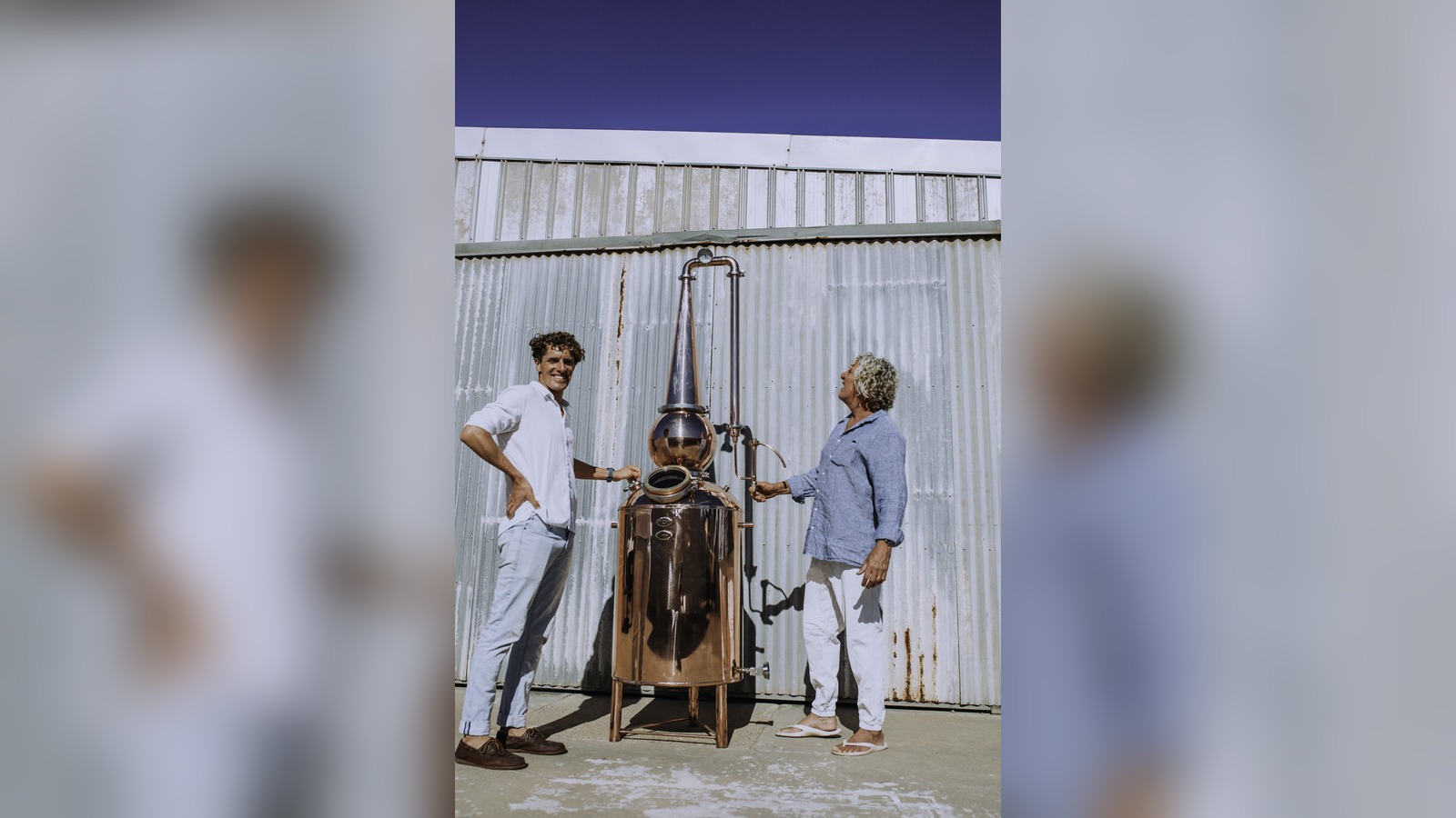 Two people standing either side of a copper still in front of a corrugated iron wall.