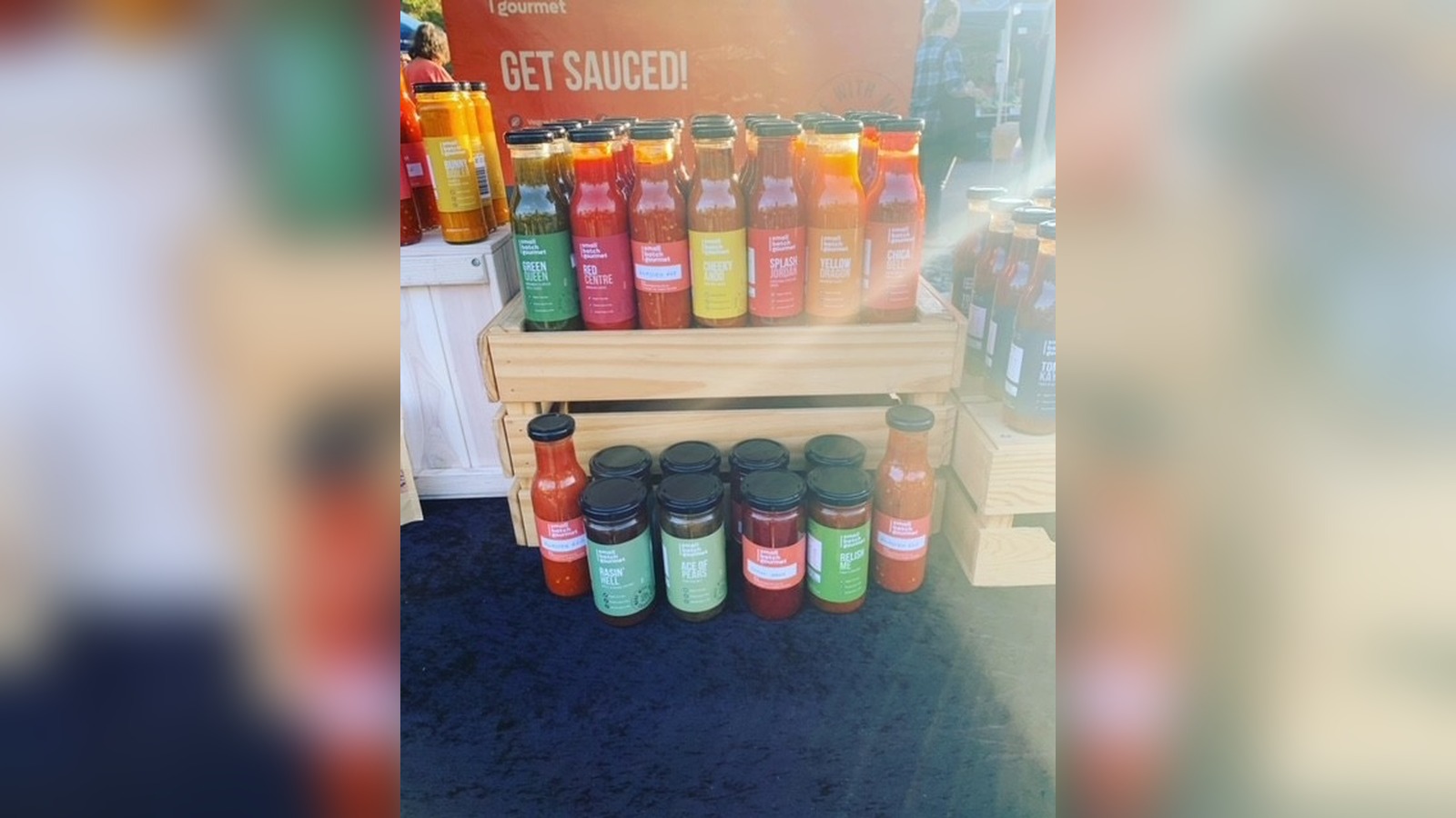 Rows of Small Batch Gourmet sauce bottles stacked on a wooden crate beneath a "Get Sauced!" banner.